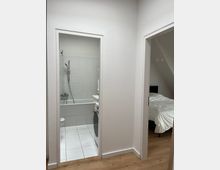 Ein schlichter Innenraum mit Blick auf zwei Türen: links ein kleines weiß gefliestes Badezimmer mit Badewanne, Duscharmatur und Waschmaschine, rechts ein Schlafzimmer mit einem weißen Bett und einer schrägen Zimmerdecke. Der Boden besteht aus hellem Holz.