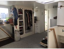 Innenraum eines kleinen Wohnbereichs mit heller Einrichtung. Zu sehen sind eine schmale Küche mit Regalen und Kleidungsaufbewahrung sowie ein Tisch mit Pflanzen und Küchenutensilien. Ein großes Fenster lässt Tageslicht herein, und eine Tür führt in einen weiteren Raum.