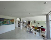 Heller Wohnraum mit offener Küche und Essbereich, hellem Laminatboden und teilweise schrägen Wänden. Die Einrichtung umfasst bunte Stühle, eine Glasplatte als Tisch, ein Regal mit Fächern, sowie ein großes, buntes Gemälde an einer weißen Wand.