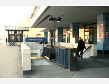 Eingang zum U-Bahnhof Rathaus Steglitz in Berlin mit Rolltreppe und Treppe, umgeben von einem modernen Gebäude mit blauen Fassadenelementen. Im Hintergrund sind Fahrradbügel, Gehweg und ein McDonald's-Schild zu erkennen.