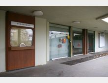 Eingangsbereich eines Gebäudes mit Glasfronten und doppelflügeligen Glastüren, die rote Griffe aufweisen. Links befindet sich ein Holzkasten mit der Aufschrift „Programme“, daneben ein überdachter Gehweg mit einem Gitterbodenbelag.