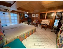 Ein gemütlich eingerichtetes Zimmer mit Holzwänden und -decke, einem Doppelbett mit buntem Rahmen, gefliesten weißen Boden und mehreren Möbeln, darunter ein Fernseher, ein Spiegel und ein kleiner Schrank mit Glastüren. Tageslicht strömt durch eine Fensterfront mit Jalousien auf der linken Seite.