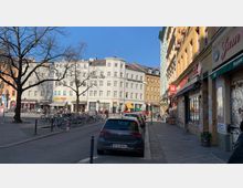 Straßenszene in einer urbanen Umgebung mit mehrstöckigen Wohn- und Geschäftshäusern; links ein großer Baum und ein Fahrradparkplatz, rechts eine Reihe von Geschäften und geparkte Autos entlang der Straße. Im Hintergrund ein helles Gebäude mit geschwungener Fassade.