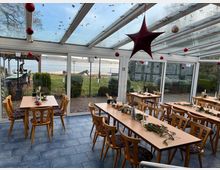 Wintergarten eines Cafés oder Restaurants mit Holztischen und -stühlen, weihnachtlicher Dekoration und Blick auf eine Rasenfläche sowie einen See im Hintergrund. Die Glasdecke und -wände lassen viel Tageslicht herein, und rote Kugeln sowie ein großer Stern hängen von der Decke.