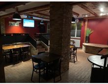 Gemütlicher Bar- oder Restaurantbereich mit Backsteinwänden und -böden, dunkelroten Wänden und Holztischen mit schwarzen Stühlen. Im Raum befinden sich Treppenstufen, eine Wand mit Fernseher und ein dekorativer Pflanzentopf in einer Ecke.