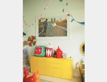 Ein bunt dekoriertes Kinderzimmer mit einer gelben Kommode, auf der Spielsachen stehen, darunter große Stoffwürfel und andere Figuren. An der Wand hängt ein gerahmtes Bild, das eine Person und einen Roboter auf einer Straße zeigt. Im Raum befinden sich farbenfrohe Dekorationen wie Girlanden und Spielzeuge.