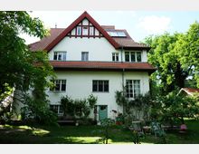 Zweistöckiges Wohnhaus mit weißer Fassade, rotem Satteldach und grüner Eingangstür, umgeben von einem Garten mit Bäumen, Sträuchern und Gartenmöbeln. Im Hintergrund sind weitere Baumbestände und ein Nebengebäude erkennbar.