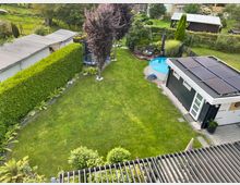 Ein gepflegter Garten mit einer zentralen Rasenfläche, umgeben von Hecken und einzelnen Bäumen. Rechts im Bild befindet sich ein Gartenhaus mit Solarpanels auf dem Dach, daneben ein runder, aufstellbarer Pool sowie eine kleine Sitzgruppe.