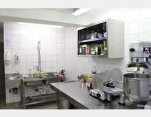 Professionelle Edelstahlküche mit Fliesenwänden, ausgestattet mit Spülbecken, Wandregalen mit Vorräten, einer Küchenmaschine, einer Aufschnittmaschine und einem Feuerlöscher.