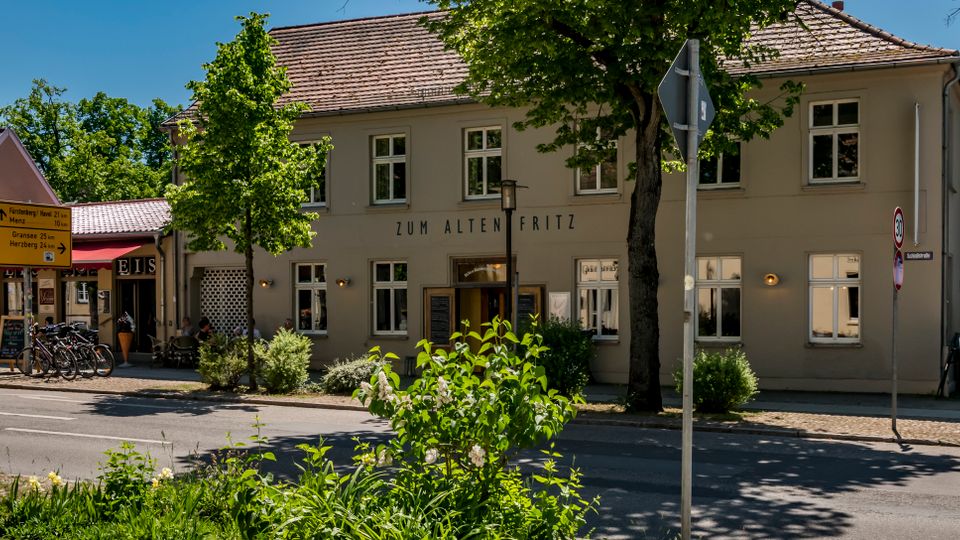 Straßenszene mit einem zweigeschossigen, hell verputzten Gasthaus mit Satteldach und der Aufschrift „Zum Alten Fritz“, umgeben von Bäumen. Vor dem Gebäude stehen Fahrräder und Straßenschilder, rechts ist eine Außengastronomie mit Tischen und Sonnenschirmen zu sehen.