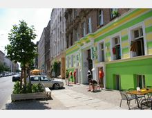 Straßenszene in einer Stadt mit einer Reihe von Altbauten, darunter ein grün-gelb gestrichenes Gebäude mit Schaufenstern und einem Ladenschild mit der Aufschrift „Skunk“. Im Vordergrund stehen ein Baum in einem Pflanzkasten, parkende Autos und ein kleiner Bereich mit Tisch und Stühlen auf dem Bürgersteig.