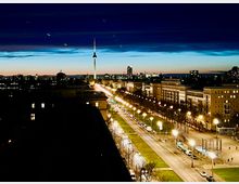 Nächtliche Stadtansicht mit dem Berliner Fernsehturm im Hintergrund, umgeben von beleuchteten Gebäuden und Straßenlaternen entlang einer breiten Allee. Der Himmel zeigt einen Verlauf von Blau zu Orange, was auf die Dämmerung hindeutet.