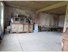 Ein rustikaler Raum mit unverputzten Ziegelwänden, einem hölzernen Buffet mit Schnitzereien und Glastüren, einigen Dekorationsgegenständen darauf, sowie einer Holzbänkecke mit grünen Metallbeinen und einem Strohgeflecht an der Decke. Der Boden besteht aus glatten, beigen Platten.