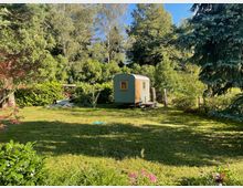 Eine grüne Wiese mit Bäumen und Büschen umgeben einen kleinen, grün gestrichenen Bauwagen mit gelben Fenstern. Im Hintergrund befinden sich weitere Vegetation und ein Stapel Feuerholz unter einem kleinen Dach.