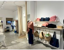 Moderner Modegeschäft-Innenraum mit gefliesten Böden, hellen Wänden und minimalistischer Gestaltung. Kleidungsstücke hängen an Kleiderständern, während gefaltete Kleidung auf Regalen liegt; Körbe und Taschen sind an einer Heizung befestigt.