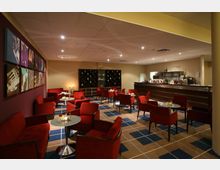 Eine moderne Hotelbar mit roten Polstersesseln und kleinen runden Tischen auf einem Teppich mit geometrischem Muster. Im Hintergrund befinden sich ein langer Tresen mit Flaschen und Gläsern sowie ein Weinregal an der Wand.