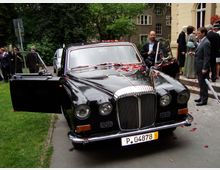 Ein klassisches schwarzes Auto steht dekoriert mit roten Rosenblättern vor einem Gebäude. Im Hintergrund sind Personen in formeller Kleidung sowie ein gepflegter Garten mit Bäumen zu sehen.