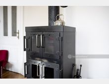 Schwarzer Kaminofen mit zwei Sichtfenstern und silbernen Griffen, darauf ein verzierter, metallischer Teekessel; im Hintergrund eine weiße Wand und eine klassische weiße Tür.