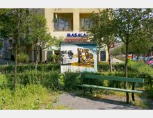 Städtische Straßenszene mit einem kleinen Parkbereich im Vordergrund, der Sitzbänke, Rasen und Bäume umfasst. Im Hintergrund ein Gebäude mit einem Restaurant namens „Masala“ und einer bemalten Wandinstallation, die ein Straßenszenen-Motiv zeigt.