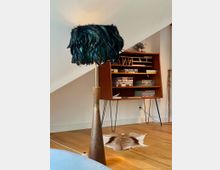 Stehlampe mit schwarzem Lampenschirm aus Federn vor einer Holzwandkonsole im Retro-Design, gefüllt mit Büchern und kleinen Gegenständen; darunter ein Kuhfell-Teppich auf Holzboden.