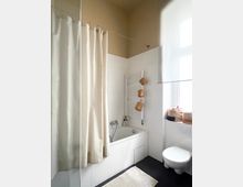 Bright bathroom with a white-tiled bathtub and shower curtain, a wall-mounted towel radiator, and a floating toilet beside a window ledge with storage baskets. Dark flooring contrasts with the white fixtures, and the ceiling is painted a warm beige.