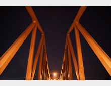 Nachtaufnahme einer modernen Brücke mit orange beleuchteten Stahlträgern, die sich symmetrisch in die Dunkelheit erstrecken. Im Hintergrund ist ein sternenklarer Himmel sichtbar.