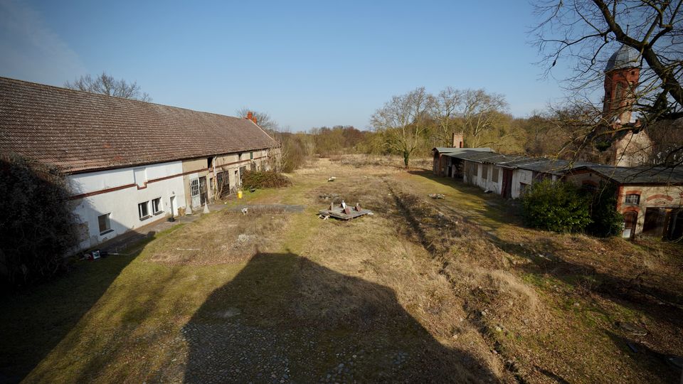Weitläufiger, verwilderter Gutshofhof mit gras- und brachbewachsenem Gelände zwischen langen, niedrigen Wirtschaftsgebäuden mit Ziegeldächern. Im Hintergrund stehen kahle Bäume, rechts ist ein runder Backsteinturm mit Kuppeldach am Rand der Anlage zu sehen.