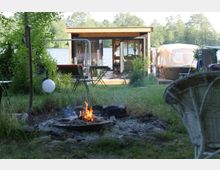 Eine kleine Holzlaube am Ufer eines Sees, umgeben von Wiesen und Bäumen, mit einem Feuerplatz im Vordergrund und einem Boot rechts daneben.