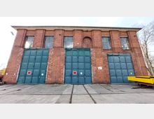 Backstein-Gebäude mit hohen, blauen Stahltoren und großen Fenstern im oberen Bereich; der Boden davor zeigt eingelassene Schienen und eine Betonfläche. Auf der rechten Seite ist ein gelbes Industriefahrzeug sichtbar.