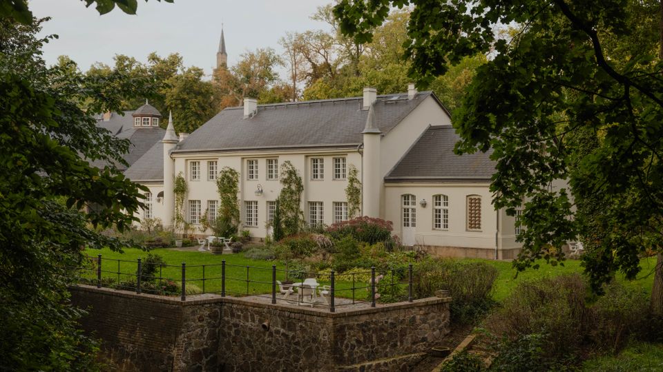 Blick auf ein helles, schlossähnliches Gebäude mit grauem Satteldach, vielen Sprossenfenstern und kleinen Türmchen, umgeben von Bäumen. Davor liegt ein gepflegter Garten mit Sitzgelegenheiten auf einer erhöhten Terrasse über einer Natursteinmauer.