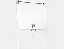 Ein minimalistisches Fotostudio mit weißem Hintergrund, einer hängenden Metallaufhängung mit Kette und einem einzelnen Holzstuhl mit schwarzem Metallgestell in der Mitte des Raumes.