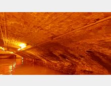 Ziegelgewölbter Kanal mit orangefarbener Beleuchtung, darunter Wasser, das die Unterseite der Struktur leicht reflektiert. Rohrleitungen verlaufen entlang der Decke.