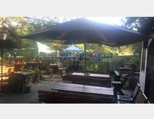 Außenterrasse eines Cafés oder Restaurants mit Holztischen und -bänken, überdacht von dunklen Sonnenschirmen. Im Hintergrund befinden sich grüne Zelte und Menschen in einem parkähnlichen Gelände mit Bäumen.