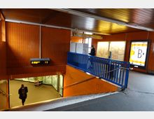 U-Bahn-Station mit orange gefliesten Wänden, blauen Geländern und einer beleuchteten Fahrgastanzeige. Im Vordergrund führt eine Treppe hinunter zu einem unteren Bahnsteig, während auf der oberen Ebene hell beleuchtete Schautafeln und Plakate sichtbar sind.