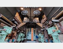 Industrieller Maschinenraum mit alten mechanischen Vorrichtungen, darunter große Zahnräder, Rohre und grün lackierte Maschinen. Der Raum hat eine Backsteinwand und eine doppelflügelige Tür im Hintergrund.