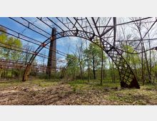 Rostige Metallkonstruktion eines verlassenen Industriegebäudes, umgeben von Bäumen und überwachsener Vegetation; im Hintergrund ist ein hoher gemauerter Schornstein zu sehen.