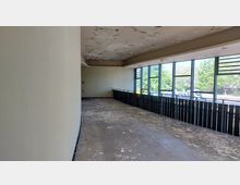 Ein leerer, verlassener Innenraum mit einer abblätternden Decke und einem Boden mit Farbresten. Große Fenster auf der rechten Seite lassen Tageslicht herein und bieten Blick auf Bäume und eine Straße draußen.