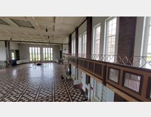 Großer Innenraum mit hohen Decken und mehreren großen Fenstern, die viel Tageslicht hereinlassen. Der Raum hat einen Boden mit geometrischen Mustern, Backsteinwände, eine Galerie aus Holz und Metall sowie einen offenen Bereich, in dem Möbel und Gegenstände verteilt sind.