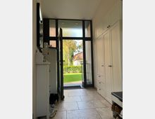 Heller Eingangsbereich mit cremefarbenem Fliesenboden, weißen Einbauschränken auf der rechten Seite und einer offenen Glastür, die zu einem Garten mit Rasen und Hecke führt. Durch große Fenster über der Tür fällt Tageslicht ein.