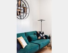 Ein modernes Wohnzimmer mit einem grünen Sofa, dekoriert mit gemusterten Kissen in Schwarz, Gold und Grün. An der weißen Wand hängt ein rundes, schwarzes Regal mit Holz-Elementen, dekoriert mit kleinen Pflanzen und Objekten. Daneben steht eine schlichte schwarze Stehleuchte.