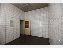 Ein leerer Raum mit weißen Wandfliesen, einem dunklen Boden und einer cremefarbenen Tür, die leicht geöffnet ist. Die Decke besteht aus grauem Beton und an den Wänden sind Steckdosen und ein kleiner Spiegel erkennbar.