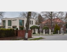 Straßenszene mit zwei Gebäuden: links ein modernes, eckiges Haus mit großen Glasfenstern und heller Fassade, rechts ein historisches Gebäude aus roten Ziegelsteinen mit rotem Walmdach. Im Vordergrund eine Straße, Bäume und ein Straßenschild.