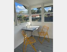 Ein kleiner, heller Wintergarten mit weißen Wänden und großen Fenstern, durch die ein Garten mit Bäumen und Häusern mit roten Dächern zu sehen ist. Im Raum stehen ein weißer Tisch und zwei gelbe Metallstühle auf einem grauen Holzboden.