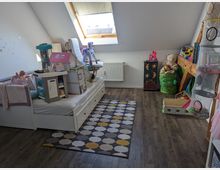 Ein Kinderzimmer mit Dachschräge, einer Dachfensteröffnung, hellem Holzboden und einer farbigen Spielzeugmatte. Das Zimmer enthält ein weißes Bett mit Spielzeugküche und Puppenhaus, Regale mit Büchern und Aufbewahrungsnetzen sowie eine Kiste mit Kuscheltieren.