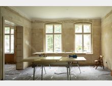 Unrenovierter Innenraum mit freigelegten, rauen Wänden und zwei großen, weiß gerahmten Sprossenfenstern, durch die Tageslicht einfällt. Im Raum stehen mehrere Arbeitstische mit Platten und Werkzeug, daneben ein Durchgang zu einem weiteren Zimmer und ein einzelner Stuhl am Rand.