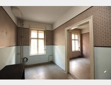 Ein leerer Raum in einem Altbau mit hohen Decken, hellen Wänden, die unten hellblau gestrichen und oben mit gemusterten Tapeten verziert sind. Zwei einfache Fenster mit Holzrahmen lassen Tageslicht herein, und ein Türdurchgang führt zu einem angrenzenden, ähnlich gestalteten Raum.