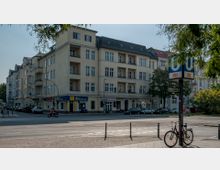 Mehrstöckiges Wohn- und Geschäftsgebäude im Stil der Gründerzeit an einer Straßenecke, mit Balkonen und Geschäften im Erdgeschoss, darunter eine Fahrschule. Im Vordergrund befindet sich ein U-Bahn-Schild der Linie U9 mit der Aufschrift "Turmstraße" sowie ein Fahrrad, das an einem Poller angeschlossen ist.