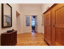 Holzvertäfelter Flur in einem Wohnhaus mit hellem Holzboden, einem großen Holzschrank auf der rechten Seite und einer Kommode mit Spiegel links. Im Hintergrund führen Doppeltüren mit Glaseinsätzen in ein Esszimmer mit großen Fenstern.