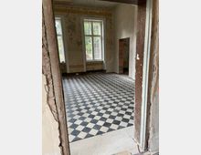 Ein unfertiger Raum mit einem schwarz-weiß karierten Fliesenboden, zwei großen Fenstern mit Sprossen und abgeblättertem Putz an den Wänden. Die Umgebung zeigt einen Renovierungszustand, mit freiliegenden Türen und unvollständigen Wandbereichen.