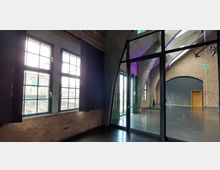 Innenraum eines historischen Backsteingebäudes mit großen Sprossenfenstern, einer Glastür, gewölbten Betondecken und einem glänzenden, dunklen Boden. Auf der rechten Seite ist eine grau gestrichene Wand mit einer hölzernen Tür zu sehen.